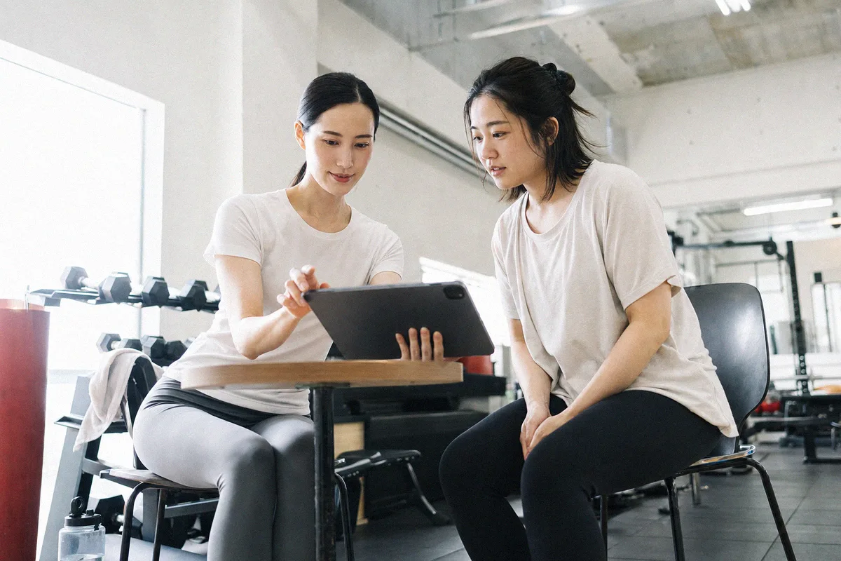 机の上のタブレットを指して内容を説明するトレーナーと、聞く利用者